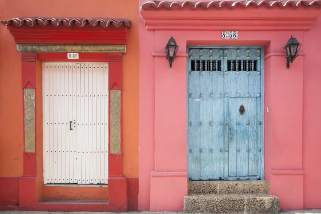 Imagen de la fachada de dos casas típicas de colombia con colores intensos en rojo y rosado junto al contraste de dos puertas pintadas de blanco y azul